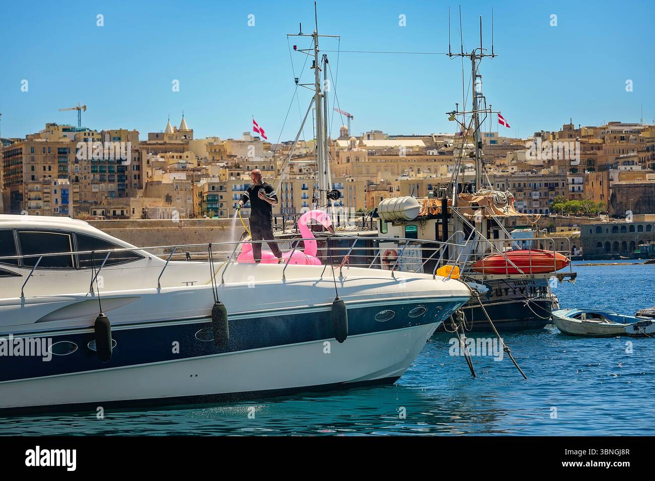 Sliema, Malte - 04.20.2025 : homme nettoyant un yacht de luxe avec un tuyau, debout à côté d'un flamant rose gonflable. Banque D'Images