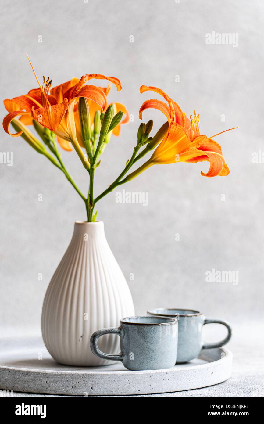 Lys tigres orangés vibrants élégamment disposés dans un vase blanc, placés sur un plateau circulaire minimaliste à côté de deux tasses en céramique sur un BA gris doux Banque D'Images