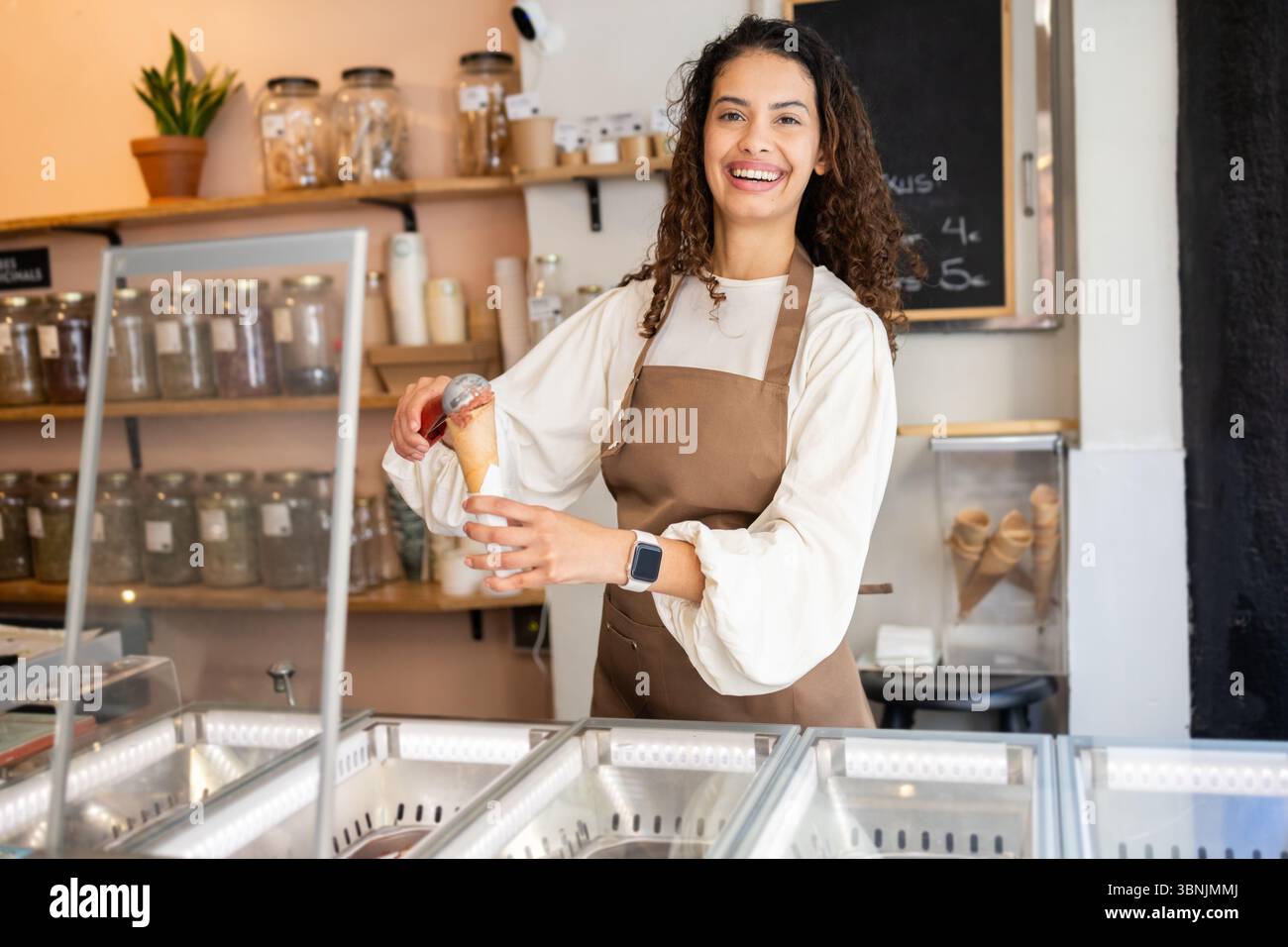 Une femme joyeuse travaillant dans un magasin de crème glacée, servant une boule de délicieuse crème glacée dans un cône. Elle porte un tablier marron et se tient derrière le comptoir Banque D'Images