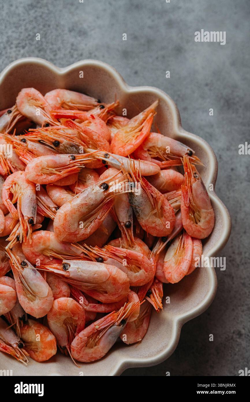 Un gros plan de crevettes roses fraîches disposées dans un bol décoratif, placées sur un fond gris texturé, soulignant leur couleur vibrante et naturelle Banque D'Images
