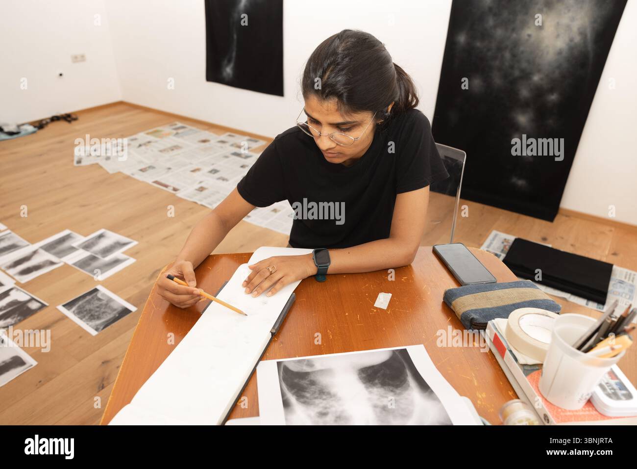 Une jeune artiste indienne engagée dans le dessin dans son studio à domicile, entourée de ses œuvres et matériaux. Elle est concentrée, portant des lunettes et un s. Banque D'Images