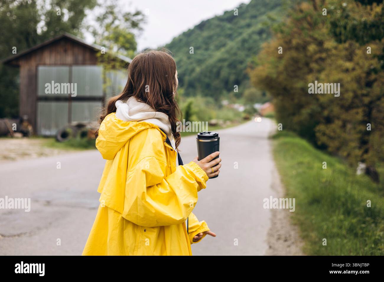 Une jeune femme se tient debout dans les montagnes sereines, portant un imperméable jaune vif, profitant de l'air frais et de la vue imprenable, tout en tenant une tasse de voyage Banque D'Images