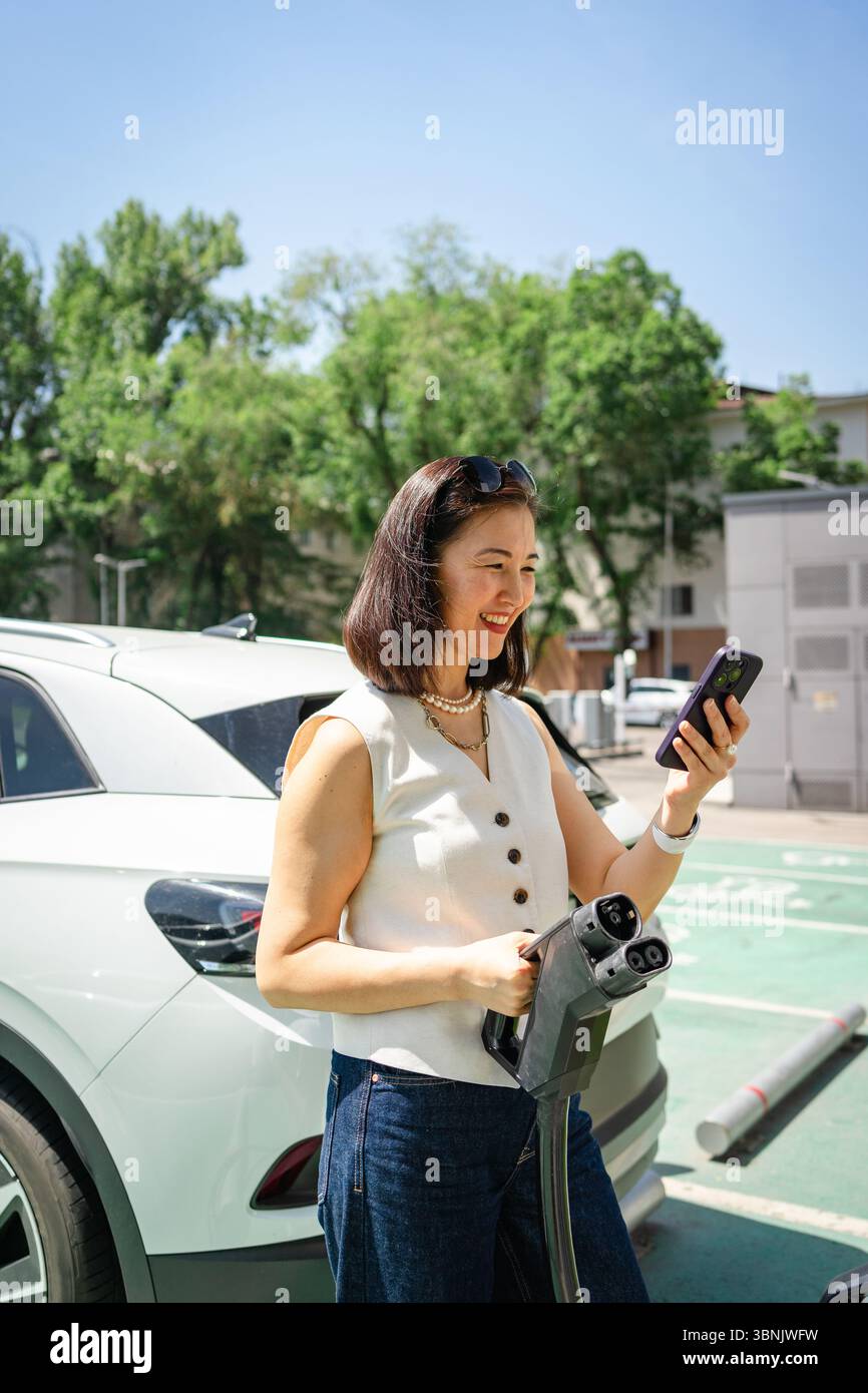 Une femme se tient près d'un véhicule électrique, tenant une prise de charge et utilisant son téléphone. Le cadre urbain vert souligne le changement écologique en urbain Banque D'Images