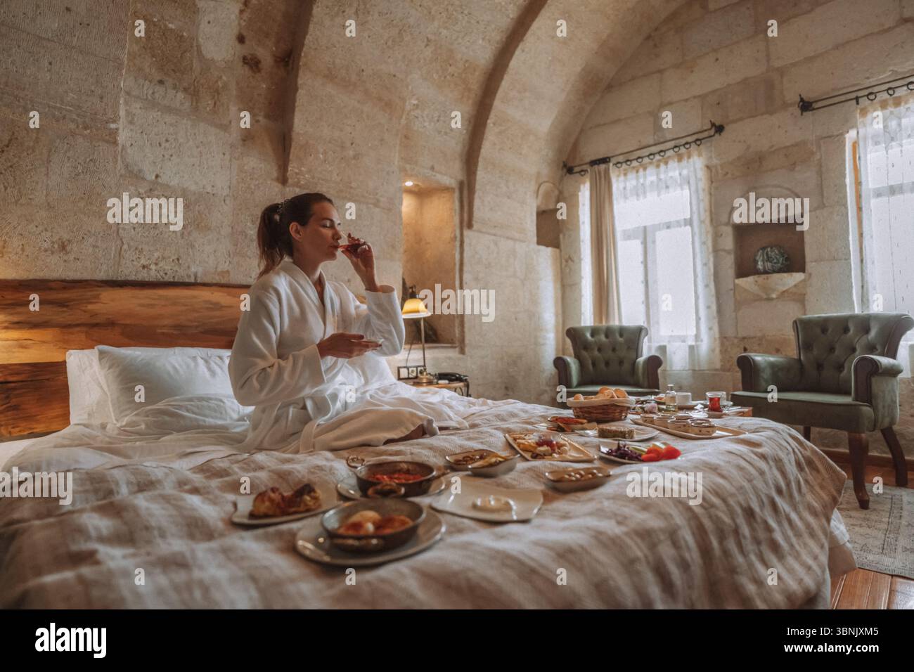 Une femme en robe blanche profite d'un copieux petit déjeuner étalé sur un lit confortable dans une chambre d'hôtel luxueuse avec d'élégants murs en pierre et une chaleur naturelle Banque D'Images