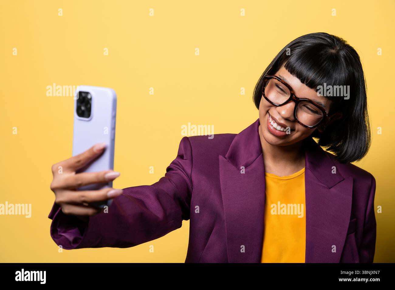 Une jeune femme hispanique dans un blazer violet sourit tout en prenant un selfie avec son smartphone sur un fond jaune vif, exsudant la confiance et Banque D'Images