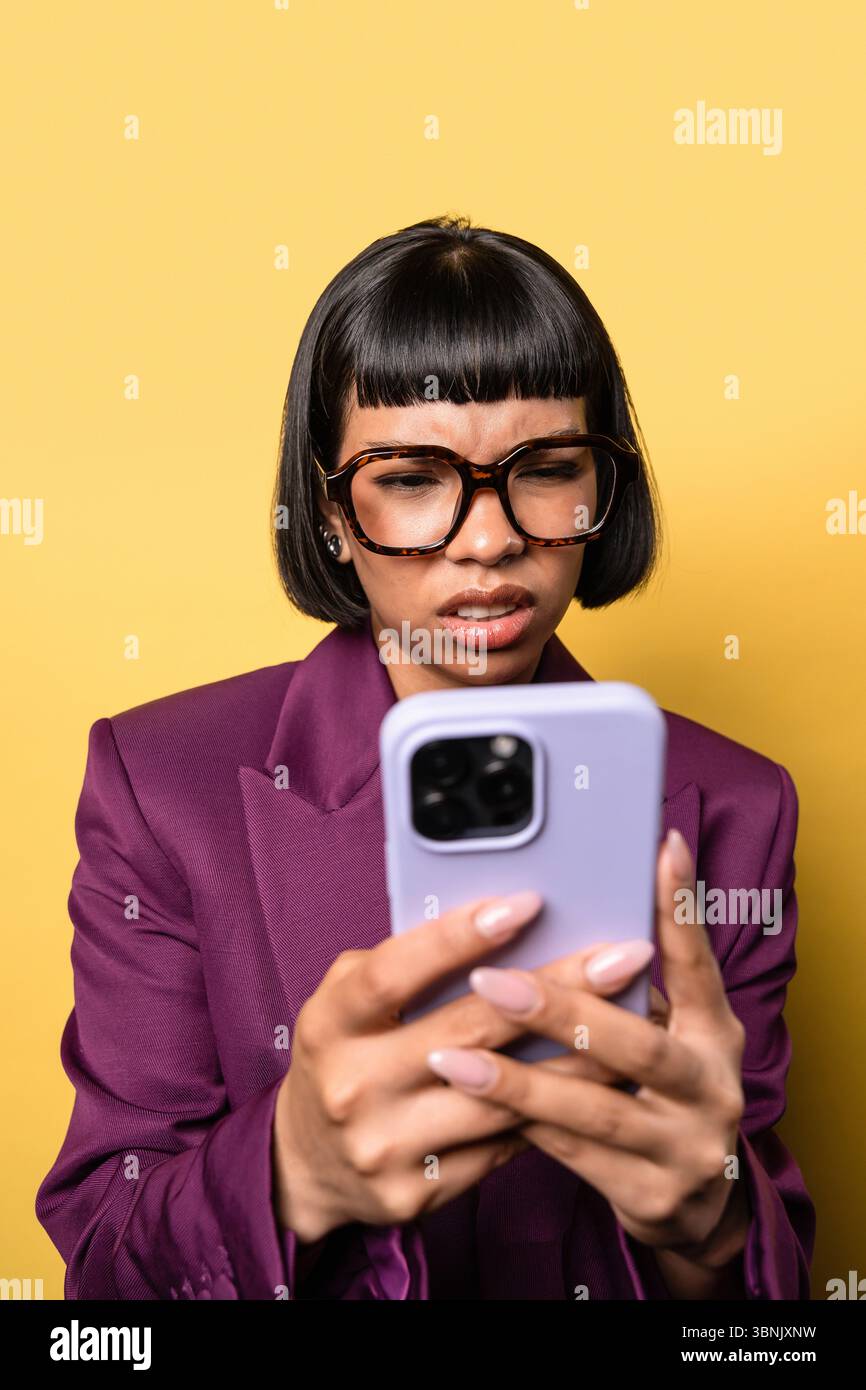Une jeune femme hispanique portant un blazer violet, aux cheveux courts et aux lunettes surdimensionnées, utilise intentionnellement son smartphone sur un fond jaune vif. Lui Banque D'Images