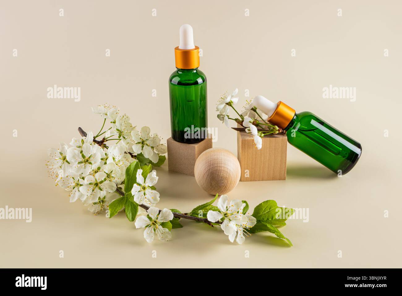 Les flacons de sérum en verre vert avec compte-gouttes dorés sont élégamment exposés aux côtés de délicates fleurs blanches et blocs de bois, créant un naturel et apaisant Banque D'Images