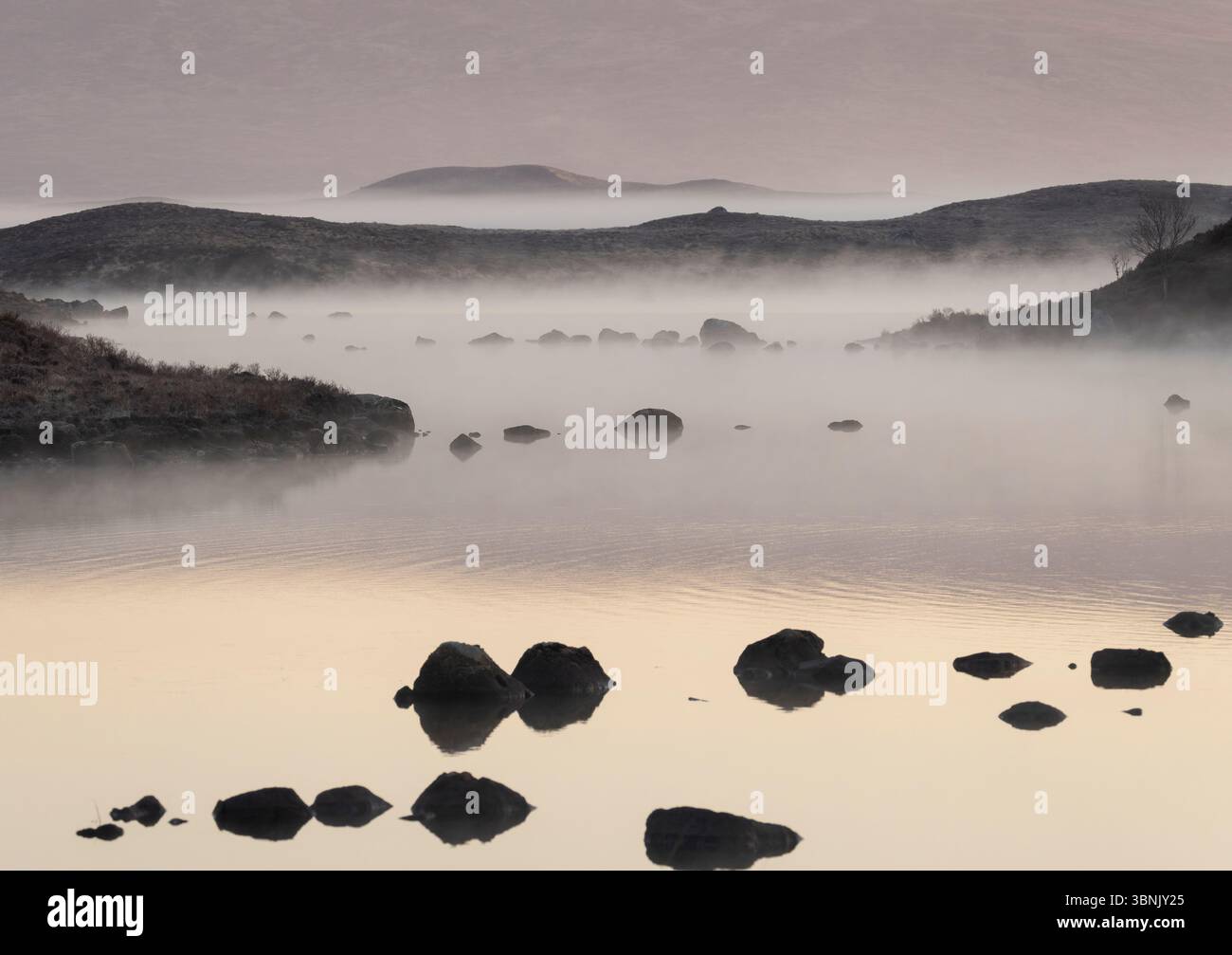 Un paysage serein de Glencoe capturé au lever du soleil, avec un lac couvert de brume parsemé de rochers et entouré de collines doucement vallonnées. Banque D'Images