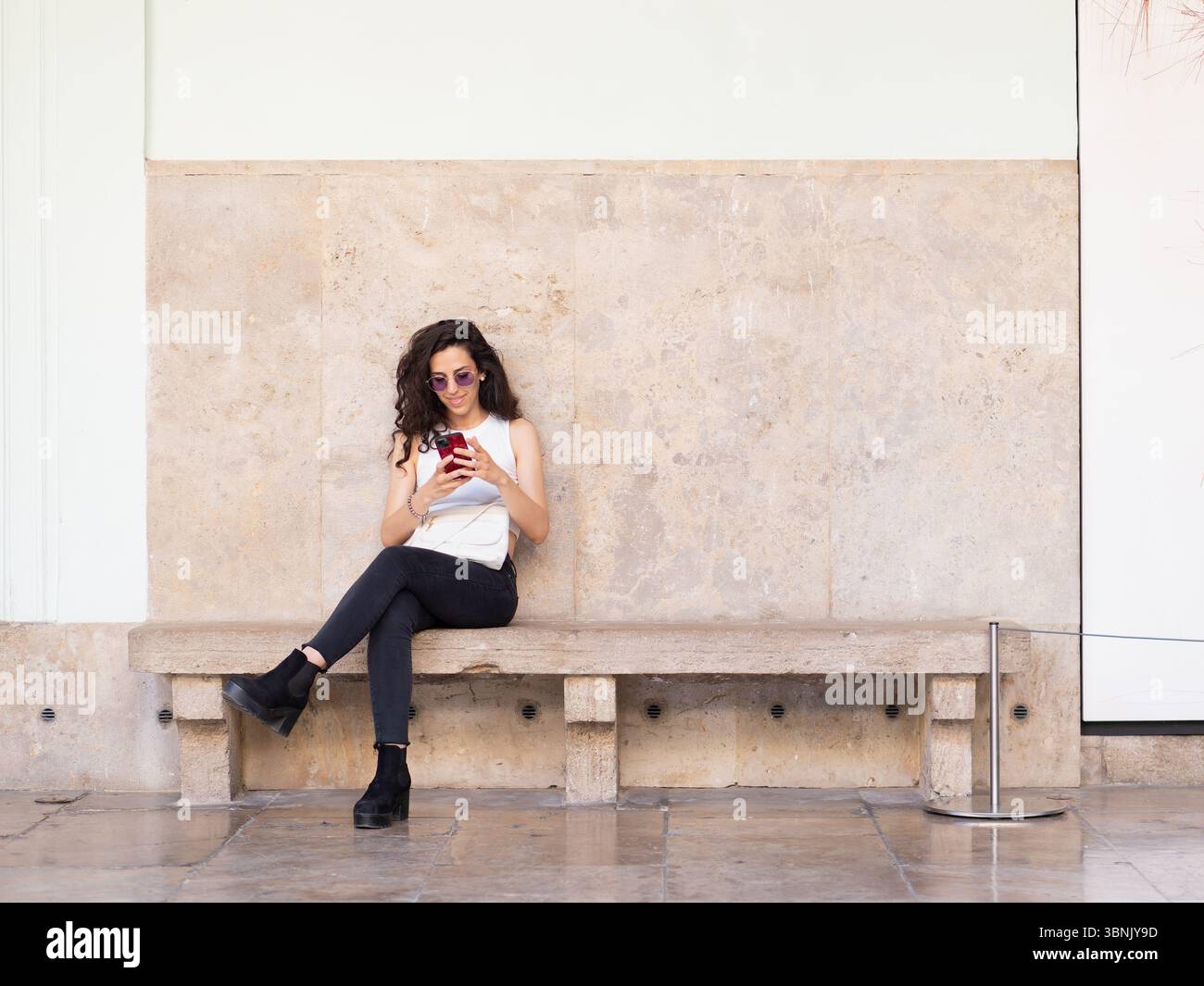 Une jeune femme en lunettes de soleil et en tenue décontractée est assise sur un banc de pierre, absorbée dans son smartphone. La toile de fond urbaine met en évidence un style de vie moderne en t Banque D'Images