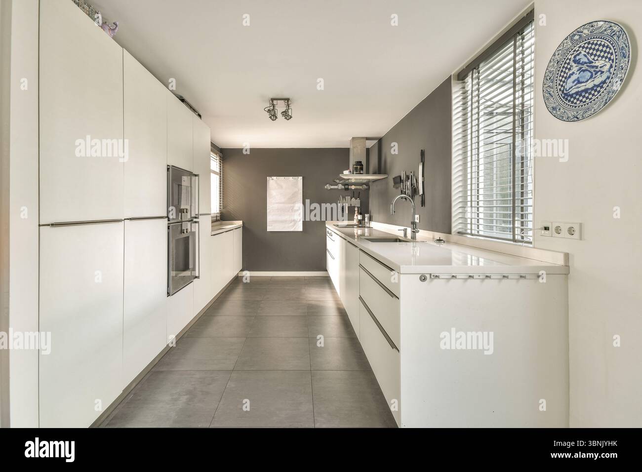 Design contemporain de la cuisine, armoires blanches élégantes et décor minimaliste. La lumière naturelle coule à travers de grandes fenêtres avec des stores, soulignant Banque D'Images