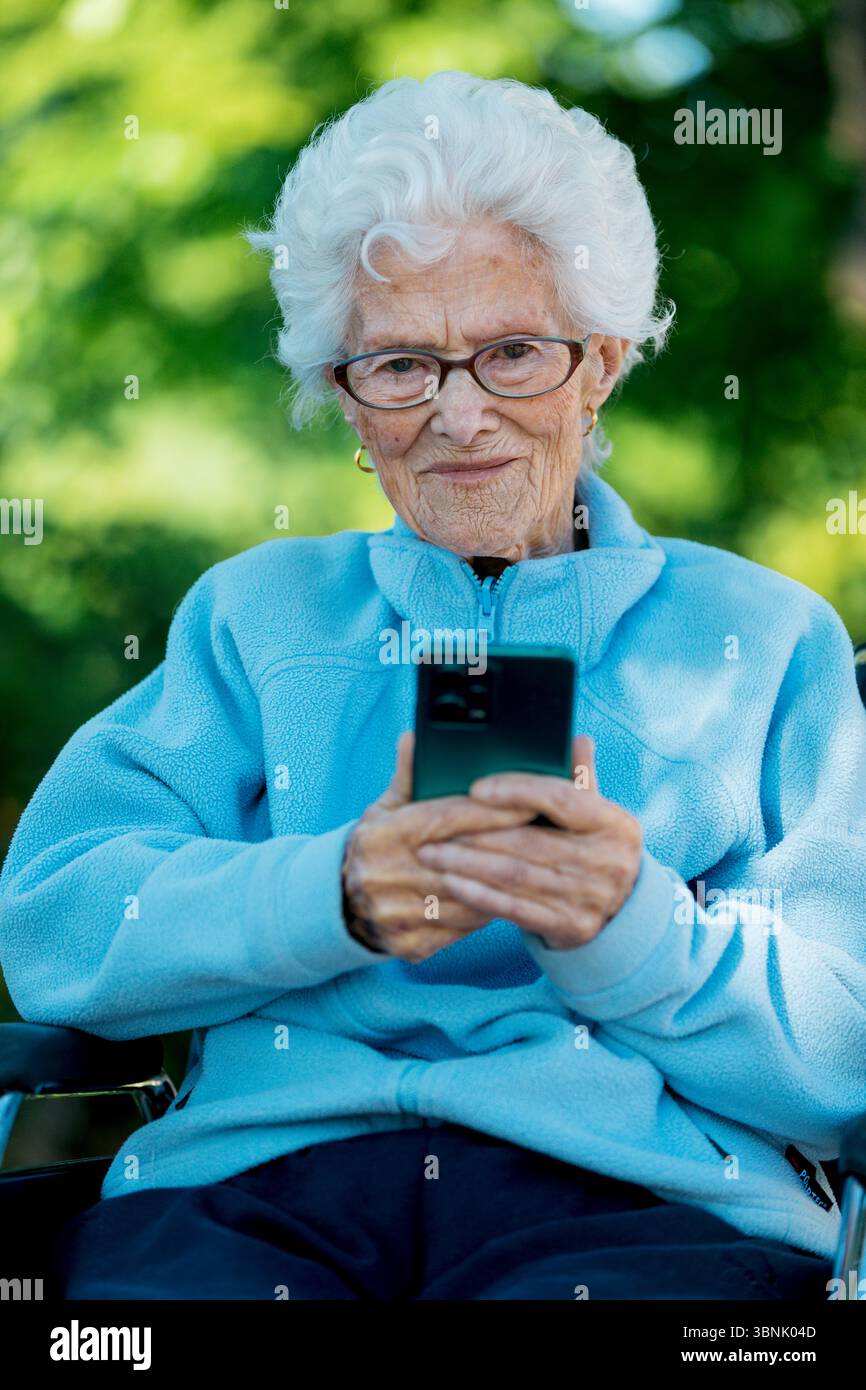 Une femme âgée dans une veste bleue, assise dans un fauteuil roulant, utilise un smartphone avec un sourire paisible. La scène est mise contre un bac extérieur vibrant Banque D'Images