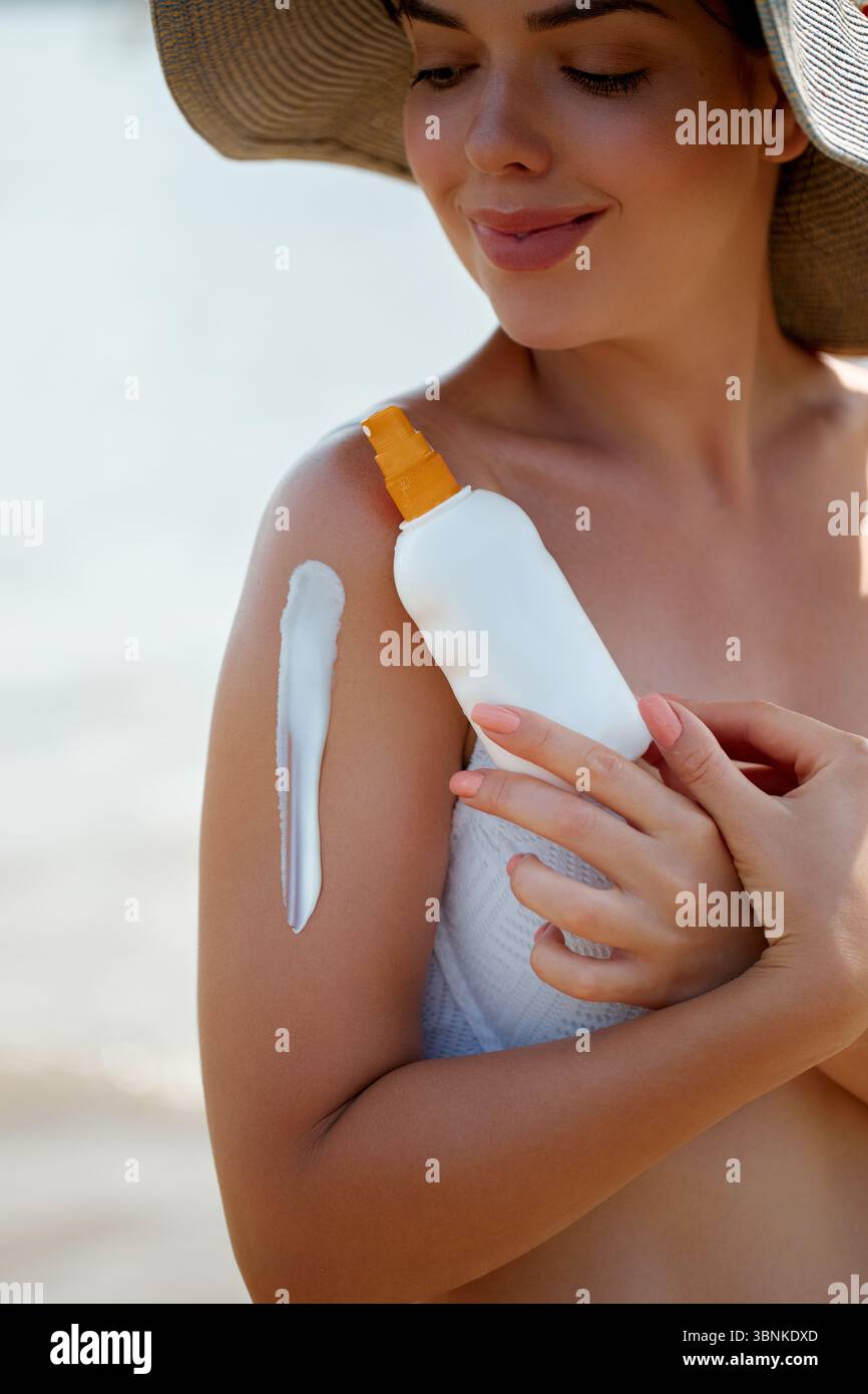 Une femme souriante tout en appliquant une lotion de crème solaire sur son épaule, profitant des vibrations ensoleillées de la plage. L'application de protection solaire met en évidence un a serein Banque D'Images