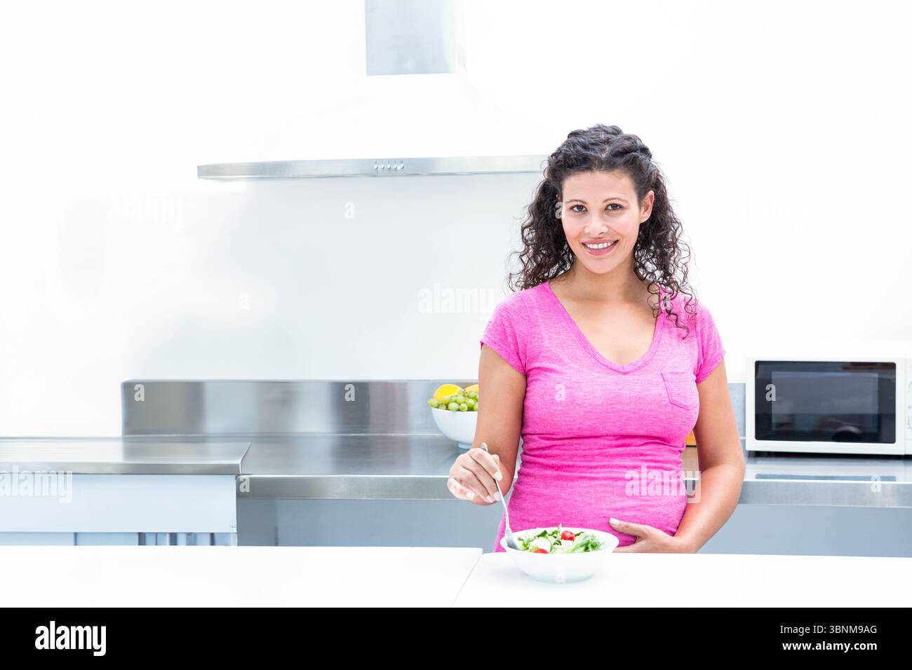 Femme enceinte debout au comptoir en acier inoxydable tenant la fourchette au-dessus du bol à salade près du bol à fruits Banque D'Images