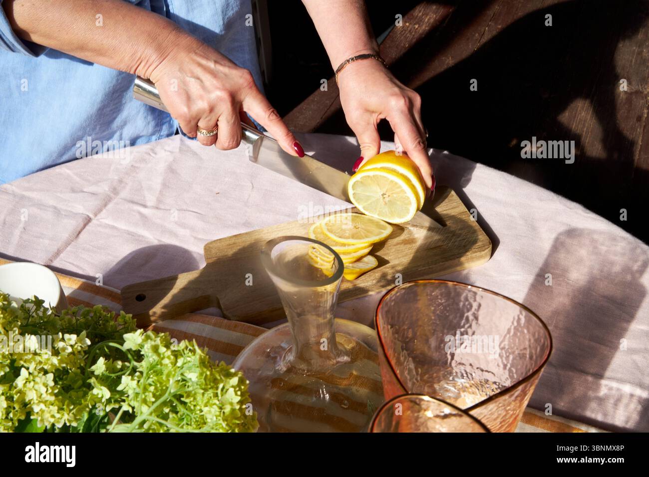 Gros plan d'une femme tranchant un citron sur une planche de bois. La lumière du soleil met en valeur les agrumes frais, la verrerie, et une ambiance estivale confortable parfaite pour la limonade ou un b Banque D'Images