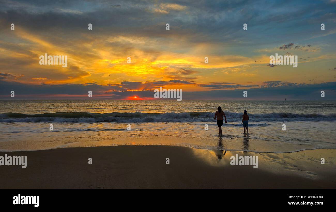 Deux adolescents pénètrent dans l'océan ou la mer lors d'un lever ou coucher de soleil pittoresque. Le soleil brille sur les nuages légers et le sable humide Banque D'Images