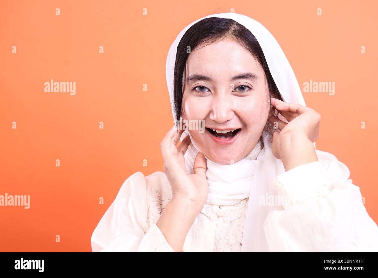 Une belle jeune femme musulmane portant un hijab blanc sourit chaleureusement à la caméra, rayonnant de positivité et de fierté culturelle, isolée sur un dos orange Banque D'Images