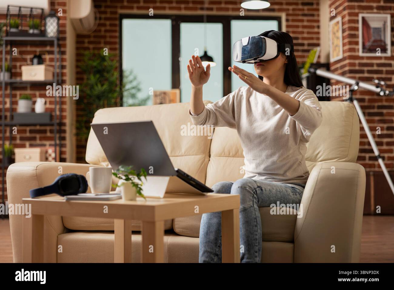 Femme d'affaires assise sur le canapé, portant un casque vr et interagissant avec l'environnement virtuel. Femme freelance explorant la technologie immersive pendant la pause dans le salon de mur de briques confortable. Banque D'Images