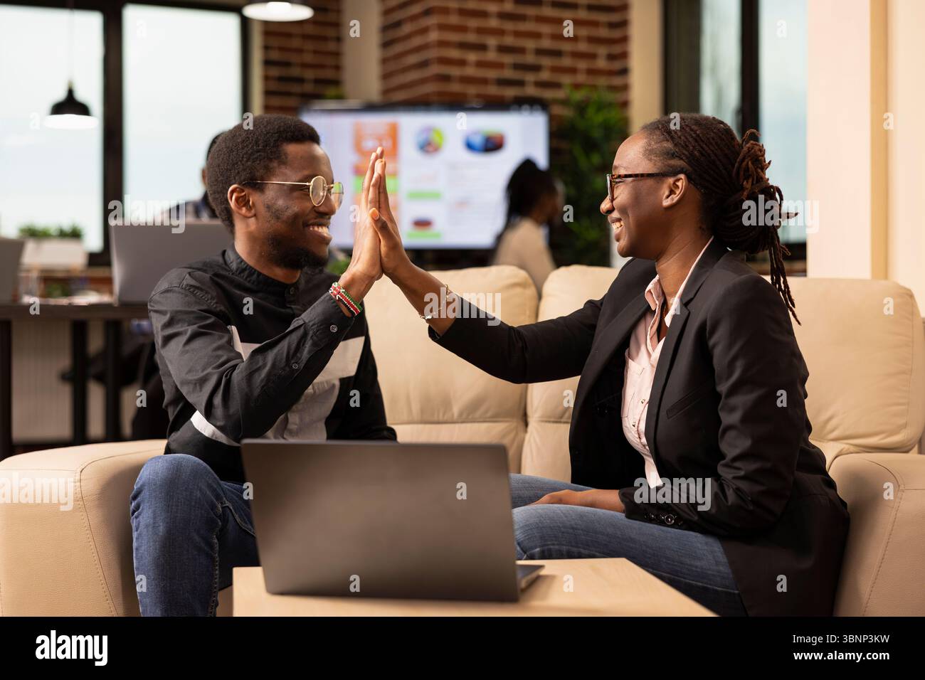 Des collègues noirs souriants échangent High Five, célébrant le travail d'équipe réussi sur un projet d'entreprise. Homme d'affaires joyeux et femme assis sur le canapé, soulignant la collaboration efficace dans le bureau de démarrage. Banque D'Images