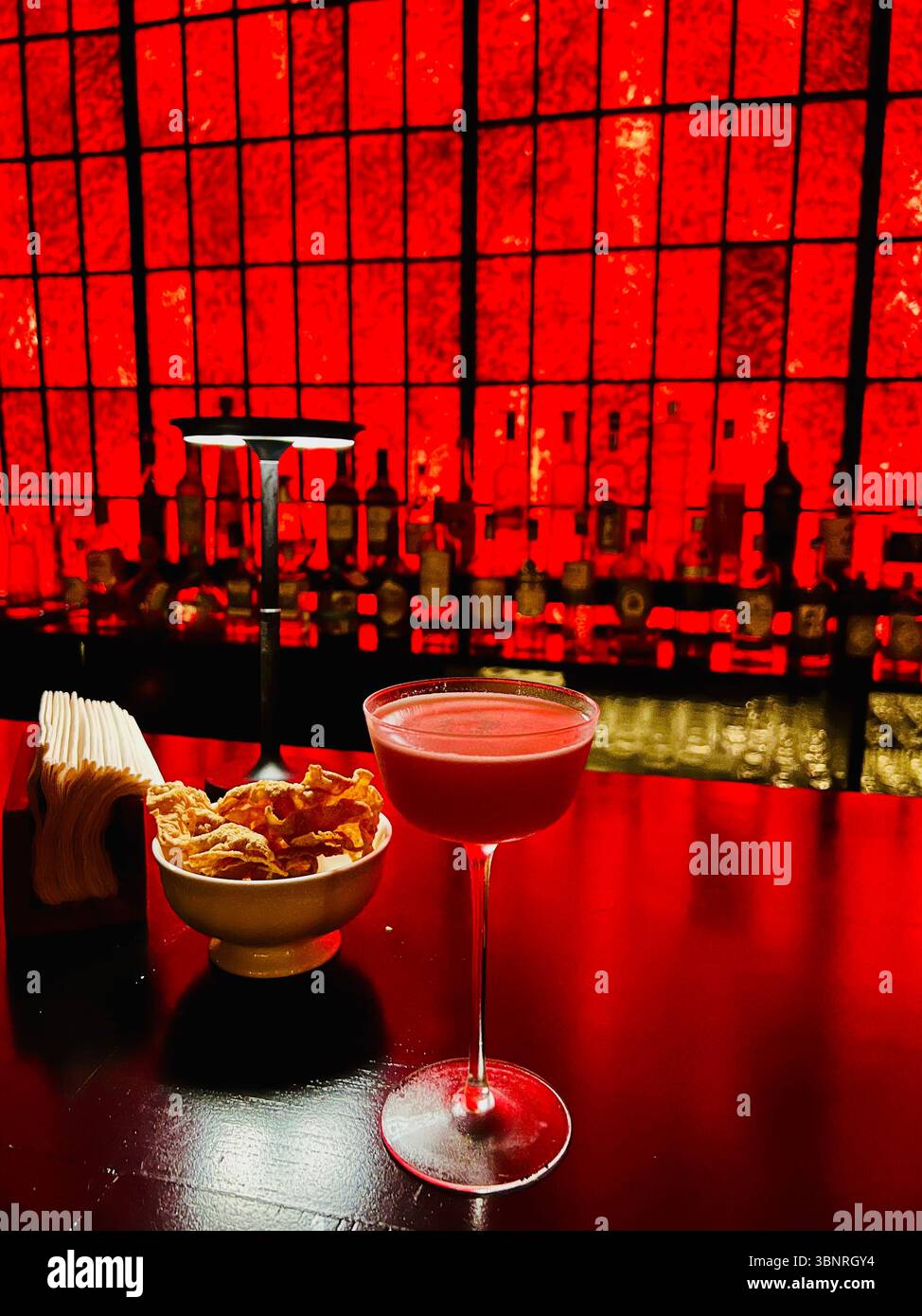 Une photo vibrante et par faible luminosité d'un cocktail rouge dans un verre à pied, accompagnée d'un bol d'en-cas croustillants, serrée sur une barre rouge étonnamment illuminée. - Image de stock capturée avec un smartphone