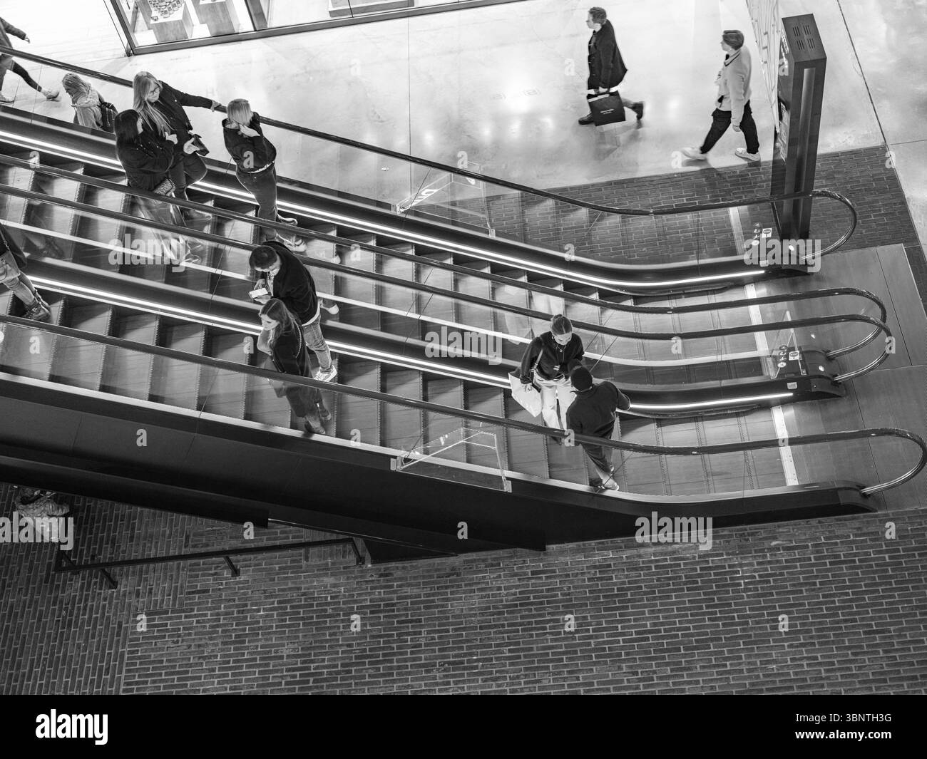 Londres, Royaume-Uni, 16 décembre 2024. Les clients empruntent les escaliers mécaniques d'un centre commercial animé du centre de Londres, vu d'en haut en noir et blanc. Banque D'Images Londres, Royaume-Uni, 16 décembre 2024. Les clients empruntent les escaliers mécaniques d'un centre commercial animé du centre de Londres, vu d'en haut en noir et blanc. Banque D'Images