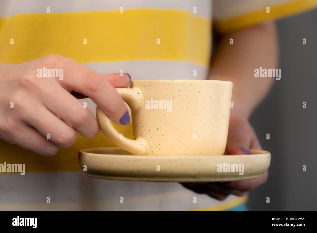 Une femme portant un T-shirt jaune et blanc avec un vernis à ongles violet tient une tasse jaune pâle et une soucoupe avec du café. Gros plan. Banque D'Images