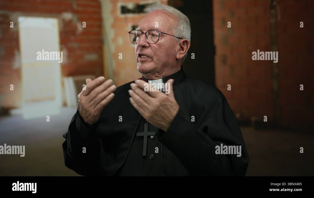 Le prêtre senior en robe noire gestes réfléchis à l'intérieur sur un chantier de construction aux murs de briques, capturant un moment de contemplation et de dévouement dans un Banque D'Images