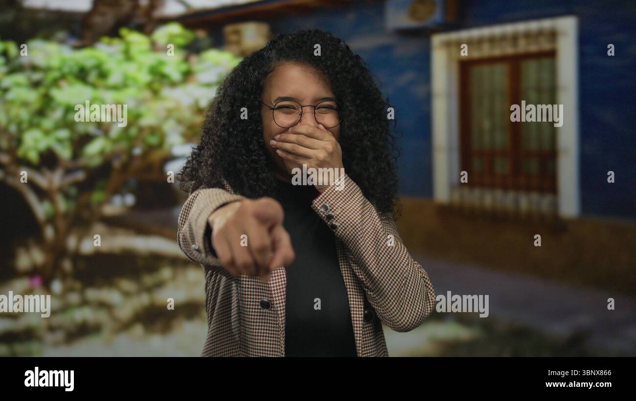 Femme riant à l'extérieur pointe ludique à la caméra avec des cheveux bouclés vibrants dans une veste élégante sur un fond coloré soulignant nat joyeux Banque D'Images