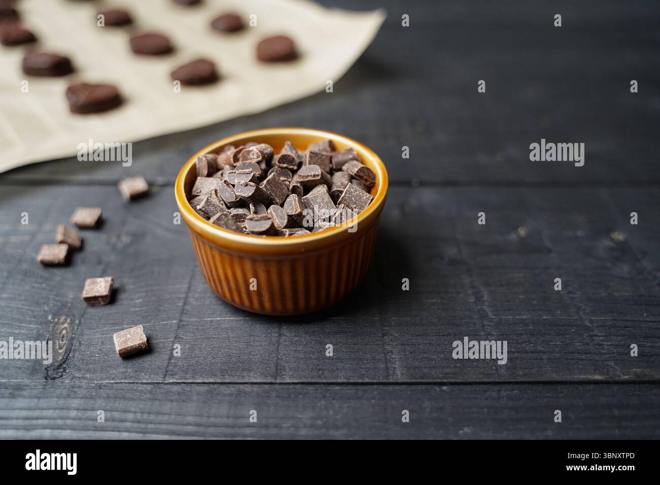 Vue de face de la pâte à biscuits aux pépites de chocolat sur papier cuisson avec bol brun de pépites de chocolat sur une surface en bois noir prête à cuire Banque D'Images