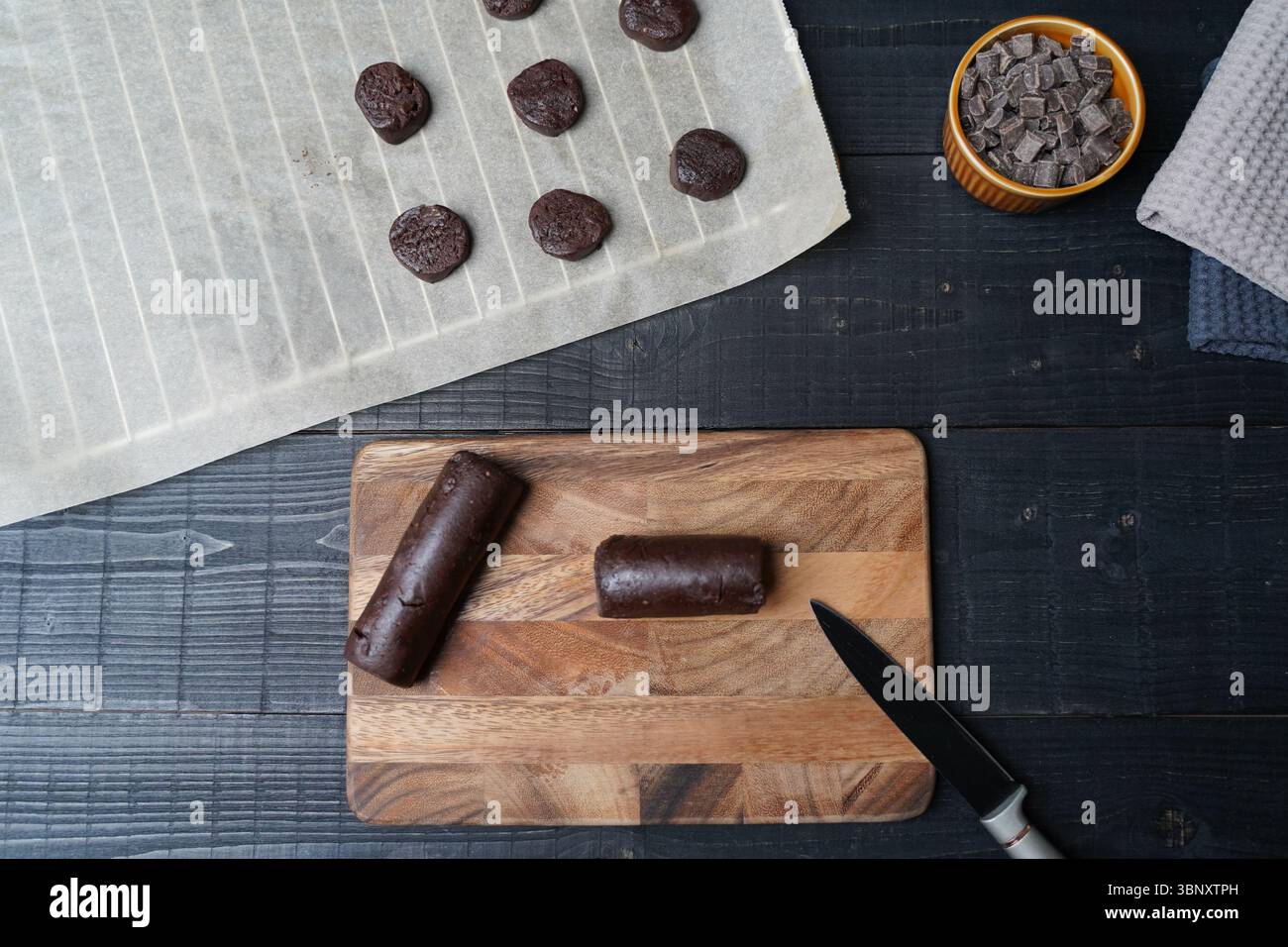 Pose plate de barre de pâte à biscuits sur du papier cuisson avec planche à découper à couteau et bol de pépites de chocolat sur fond de bois noir prêt pour la cuisson Banque D'Images