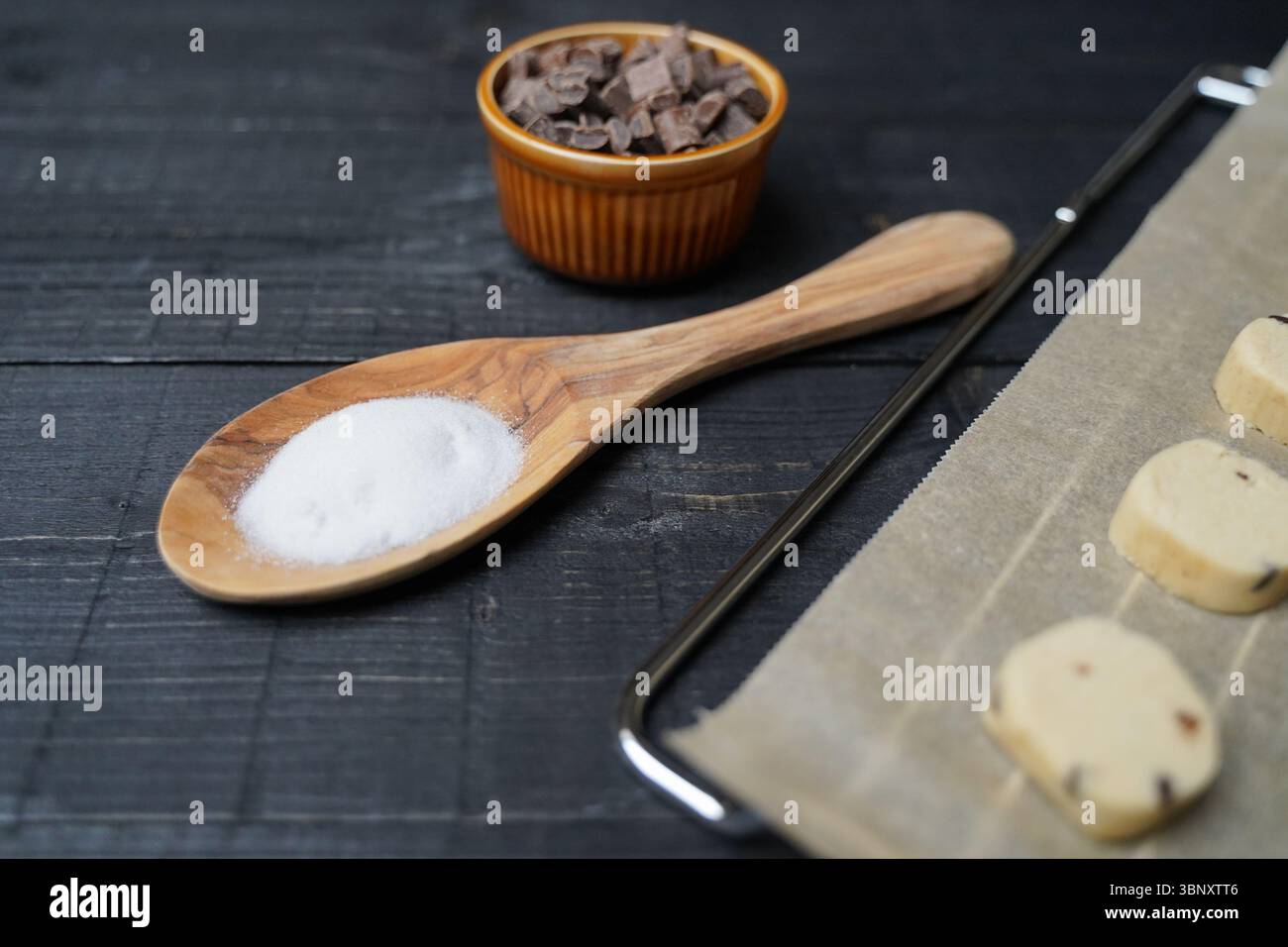Vue de dessus de la pâte à biscuits coupée sur du papier cuisson sur grille avec du sucre dans une cuillère et des pépites de chocolat dans un bol sur fond de bois noir Banque D'Images