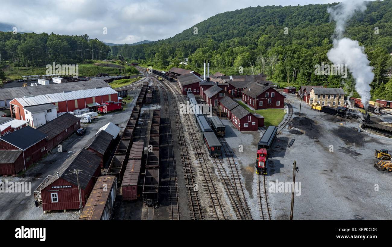 Une gare ferroviaire bien entretenue comprend des trains anciens stationnés à côté de bâtiments rustiques rouges. Des collines verdoyantes entourent la région, avec de la vapeur bouillonnante Banque D'Images