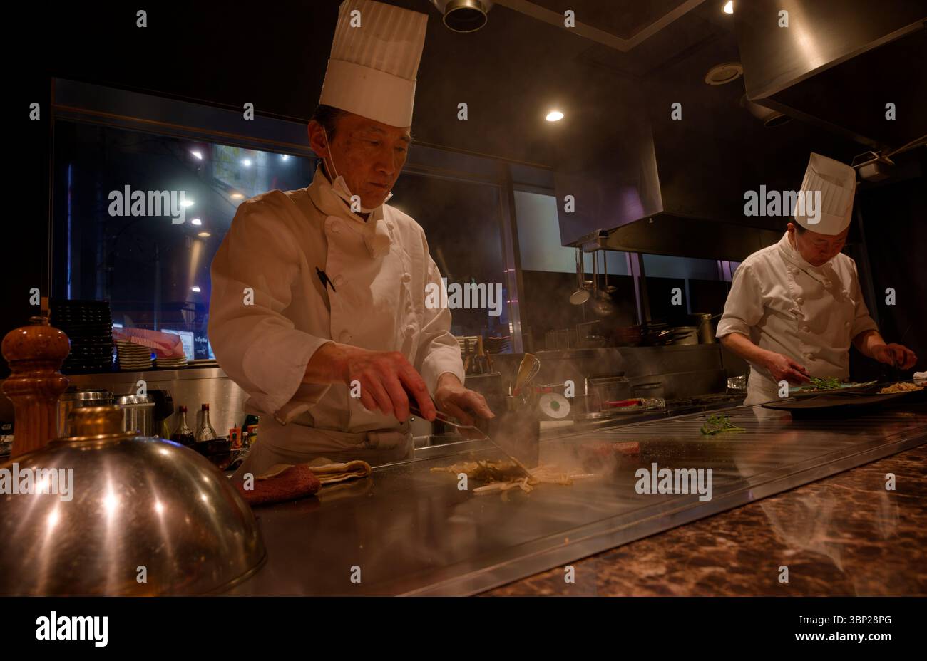 Les chefs teppanyaki préparent habilement les ingrédients sur le gril, remplissant la cuisine chaleureuse avec concentration et art culinaire Banque D'Images