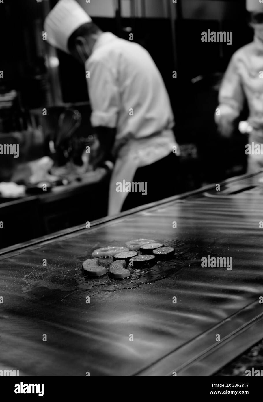 Les légumes teppanyaki grillent sur le gril chaud tandis que les chefs concentrés préparent des plats en arrière-plan Banque D'Images