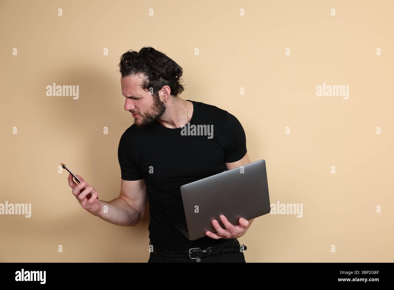 Un homme tenant un ordinateur portable regarde son smartphone avec une expression de frustration ou d'inquiétude, réagissant à son contenu, contre un dos beige Uni Banque D'Images