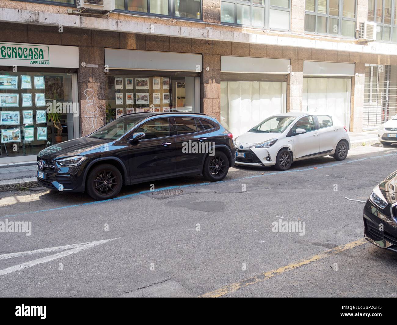 Bologne, Italie 3 juillet 2025 Noir Mercedes Benz GLA et blanc Toyota Yaris parking sur une place de parking payante devant une agence immobilière sur un urb Banque D'Images