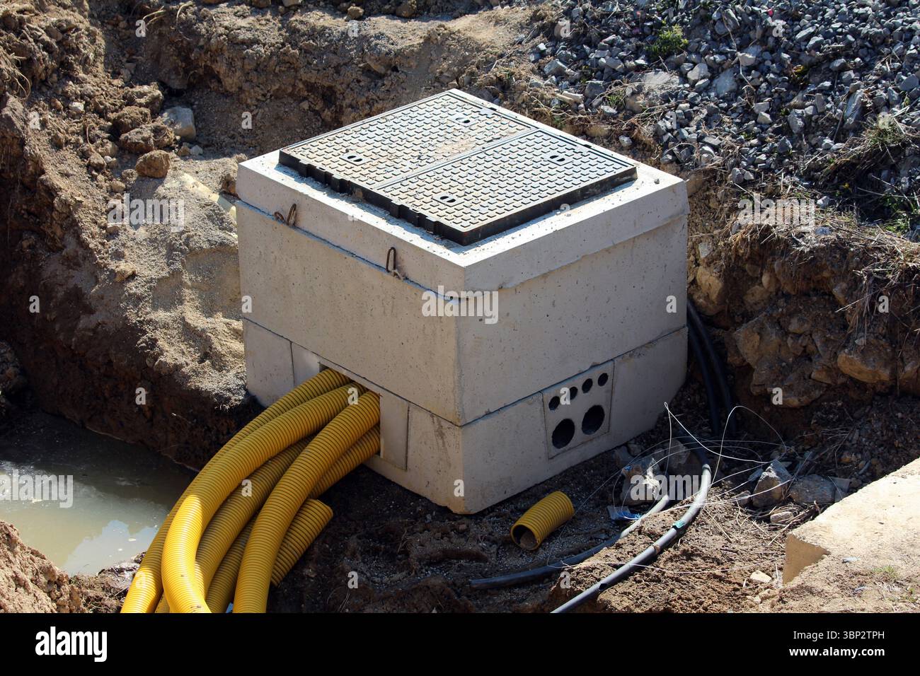 L'invention concerne une boîte de jonction de services publics en béton partiellement enterrée sur un chantier de construction, avec de multiples tuyaux de conduit ondulés jaunes pénétrant sur son côté Banque D'Images
