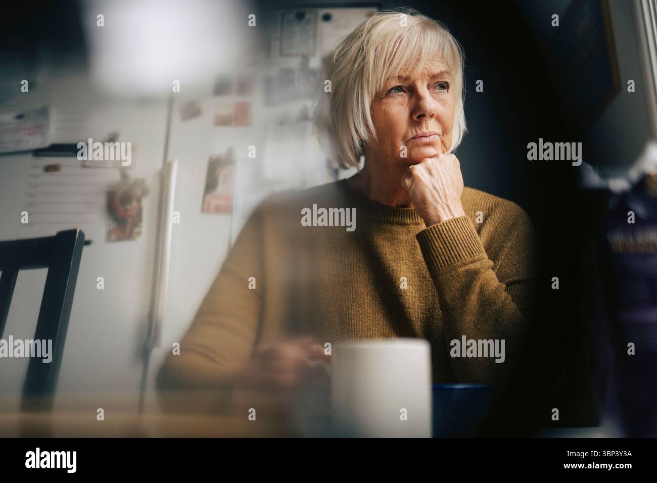 Femme senior contemplative s'appuyant sur le coude regardant loin tout en prenant un café à la maison Banque D'Images