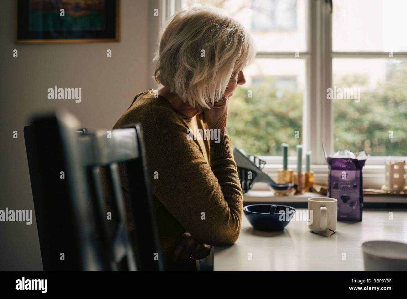 Femme âgée blonde appuyée sur le coude tout en étant assise à table dans la maison Banque D'Images