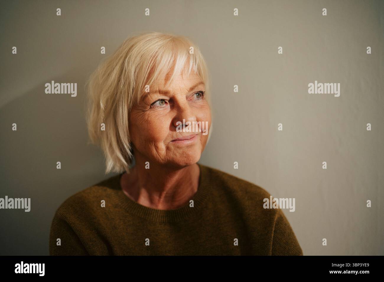 Femme âgée souriante regardant loin près du mur à la maison Banque D'Images