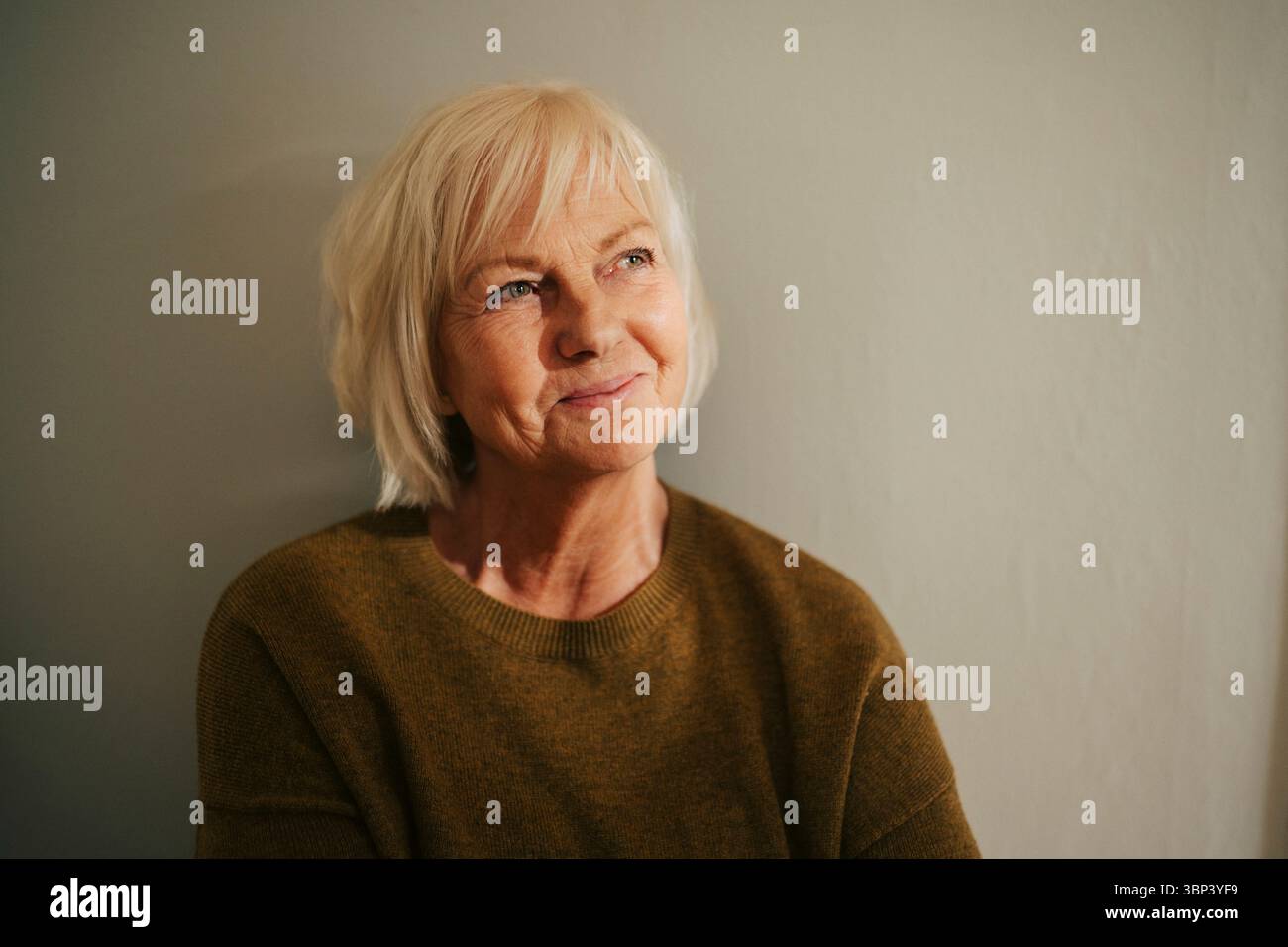 Femme âgée souriante regardant loin près du mur à la maison Banque D'Images