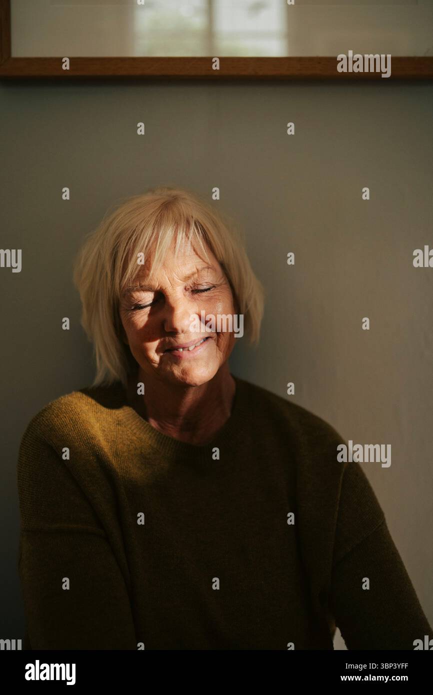 Femme âgée blonde souriante avec les yeux fermés près du mur à la maison Banque D'Images