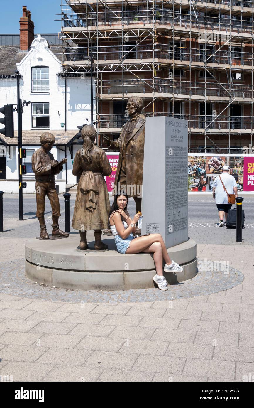 Jeune dame assise George Boole monument grignotant sur le rouleau de saucisse Greggs, St Mary's Street, Lincoln City, Lincolnshire, Angleterre, ROYAUME-UNI Banque D'Images