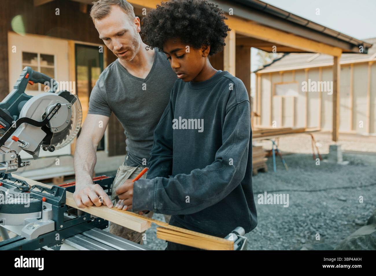 Garçon aux cheveux bouclés marquant la mesure sur une planche de bois avec l'aide du père à l'extérieur de la maison Banque D'Images