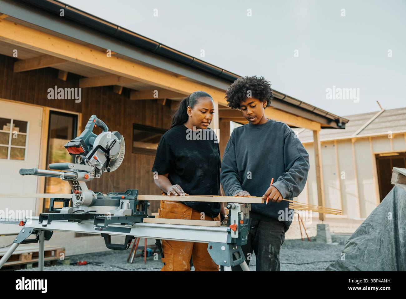 Garçon aux cheveux bouclés marquant la mesure sur une planche de bois avec l'aide de la mère à l'extérieur de la maison Banque D'Images