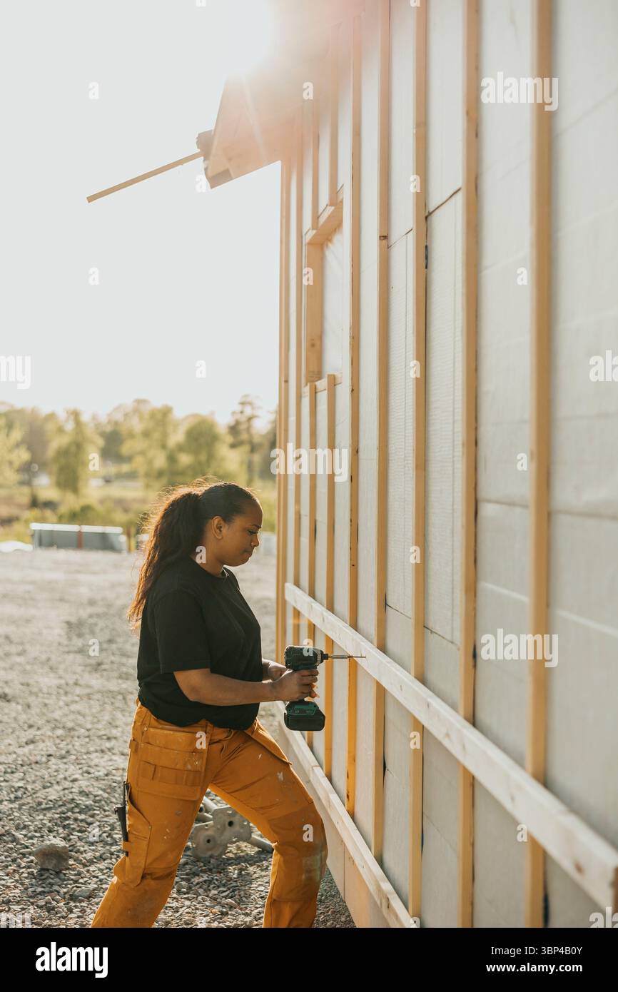 Ouvrière en utilisant la planche de bois de forage à l'extérieur de la maison au jour ensoleillé Banque D'Images