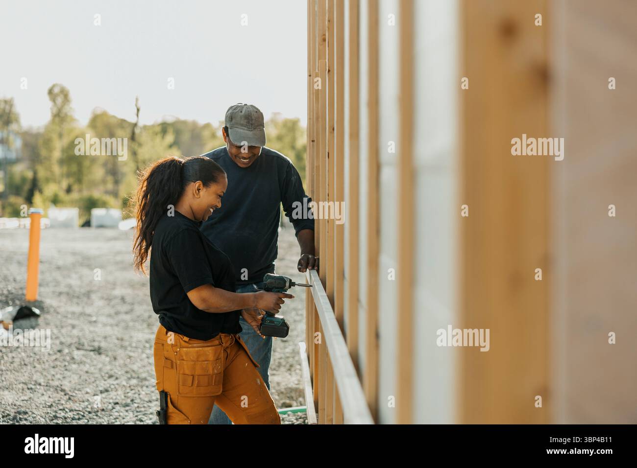 Travailleuse utilisant la perceuse sur la planche de bois debout à côté du collègue à l'extérieur de la maison Banque D'Images