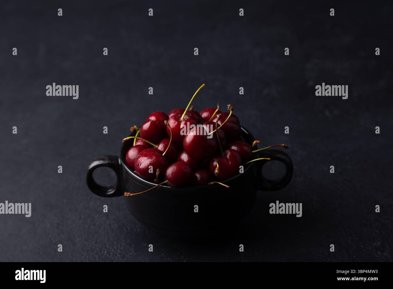 Cerises fraîches mûres juteuses dans une assiette noire sur fond noir. Fruits de saison, récolte. Mise au point sélective, gros plan. Banque D'Images