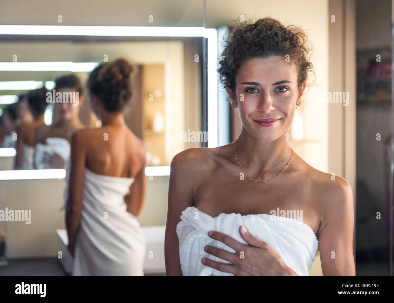 Belle femme est souriante et s'enveloppant dans une serviette blanche après un bain relaxant dans une salle de bain moderne Banque D'Images