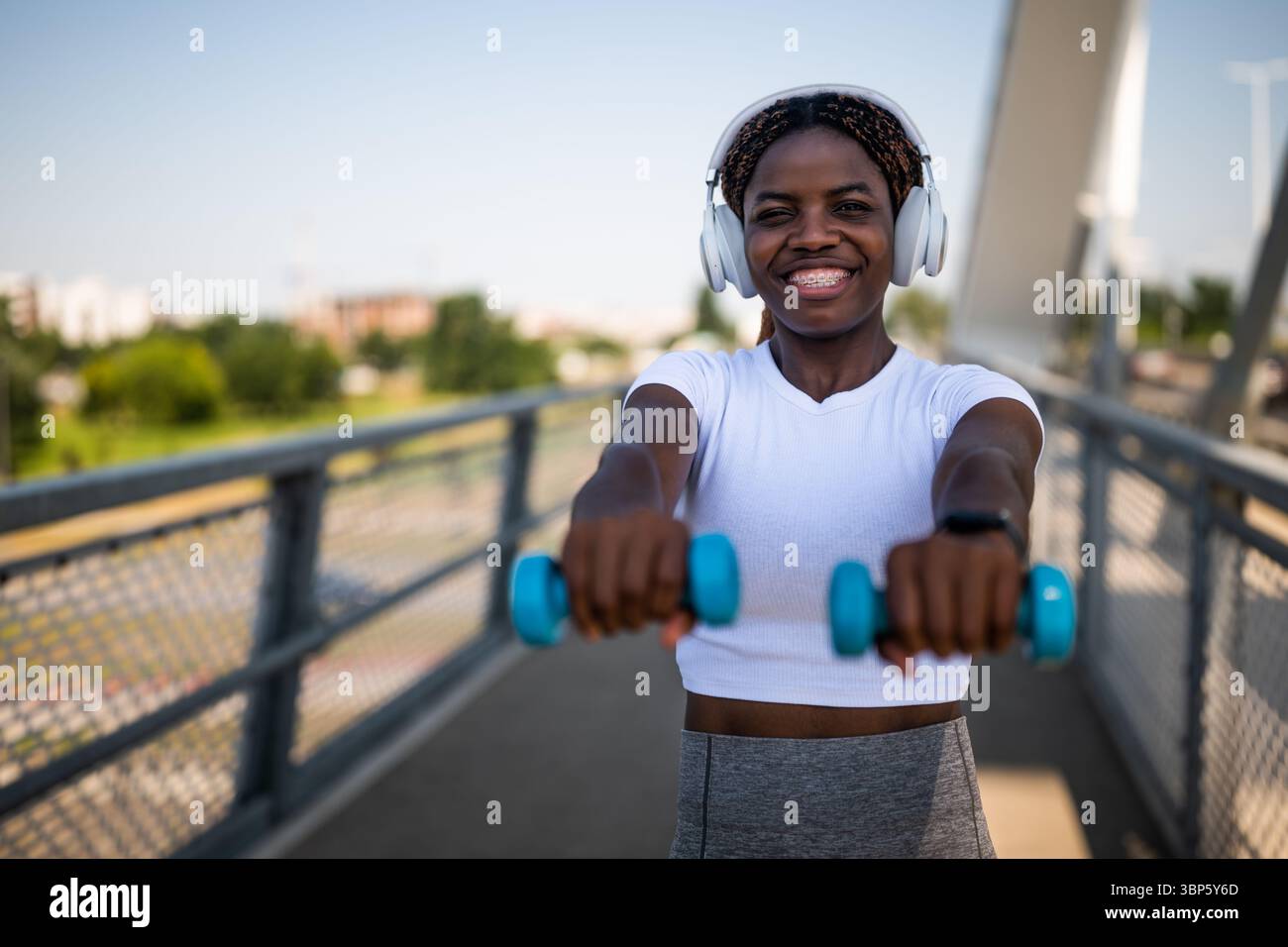 Portrait d'une jeune femme afro-américaine en vêtements de sport jouissant de l'exercice avec des poids et écoutant de la musique à travers des écouteurs sur le pont de la ville urbaine Banque D'Images