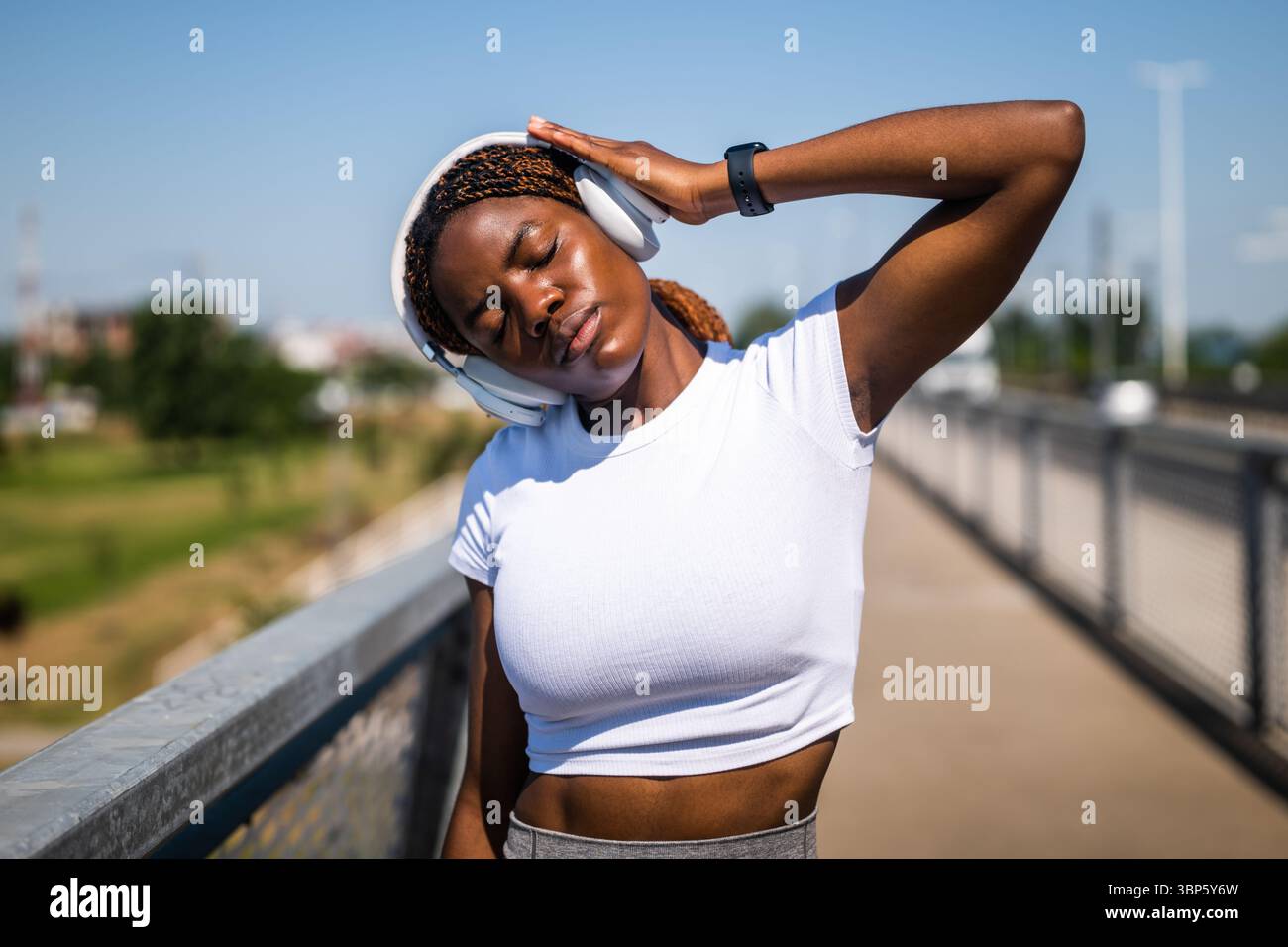 Portrait d'une jeune femme afro-américaine en vêtements de sport s'exerçant et s'étirant tout en écoutant de la musique à travers des écouteurs sur le pont de la ville. Banque D'Images